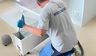 A cleaner from Housecleaners.nl kneels on the floor, deep cleaning the internal components of a radiator after removing the top grill.